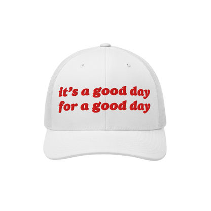 The Good Day Hat features a white trucker-style cap with a mesh back and curved brim, showcasing bright red embroidery readin