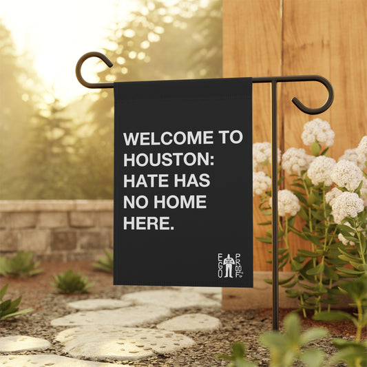 Custom Hate Has No Home Here Garden Flag displayed on a decorative metal stake beside a stone path, surrounded by greenery and white flowers, boldly states WELCOME TO HOUSTON: HATE HAS NO HOME HERE.