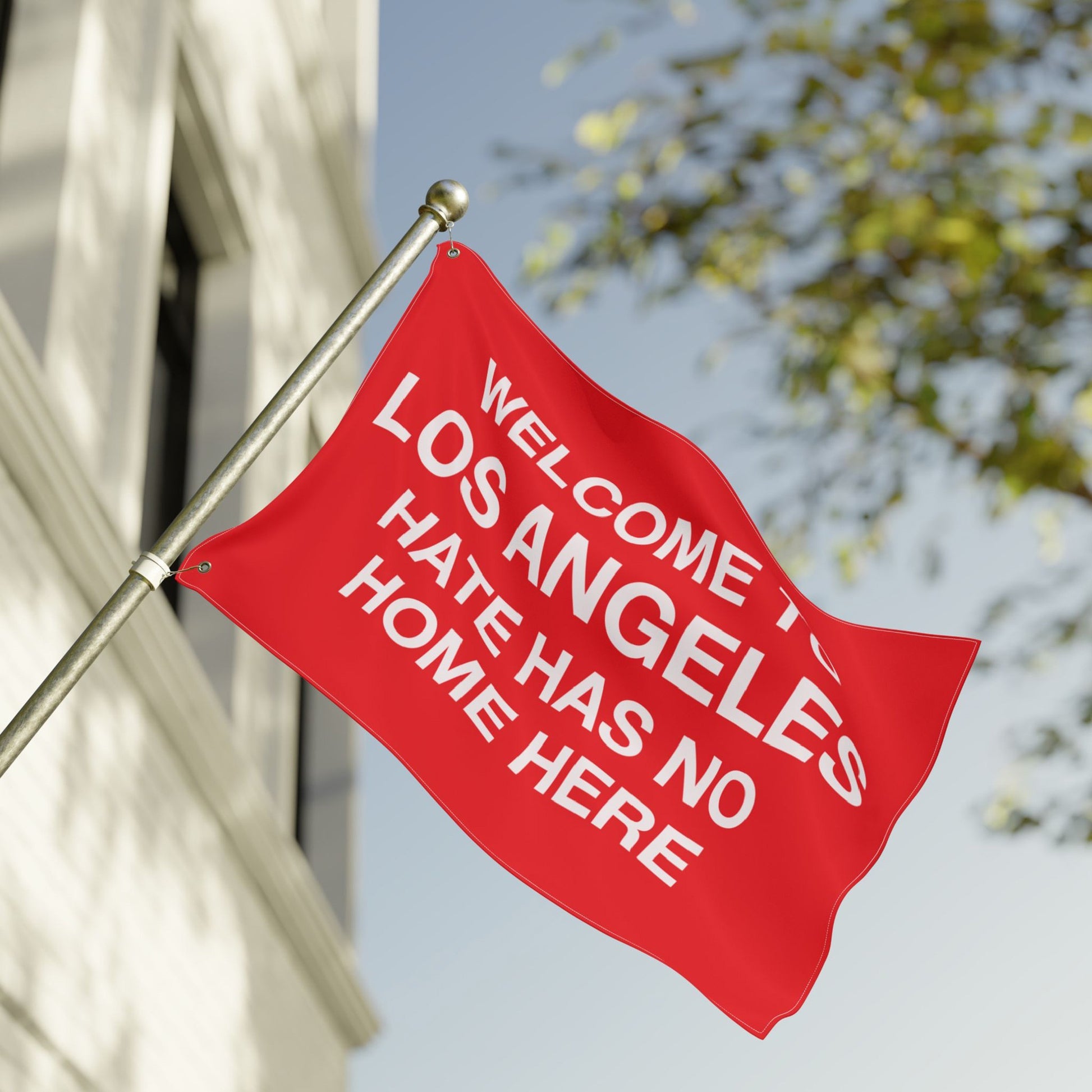 Custom Hate Has No Home Here Flag mounted on a pole, featuring bold white text Welcome to Los Angeles and Hate Has No Home Here, fluttering outside a building, symbolizing unity and acceptance.