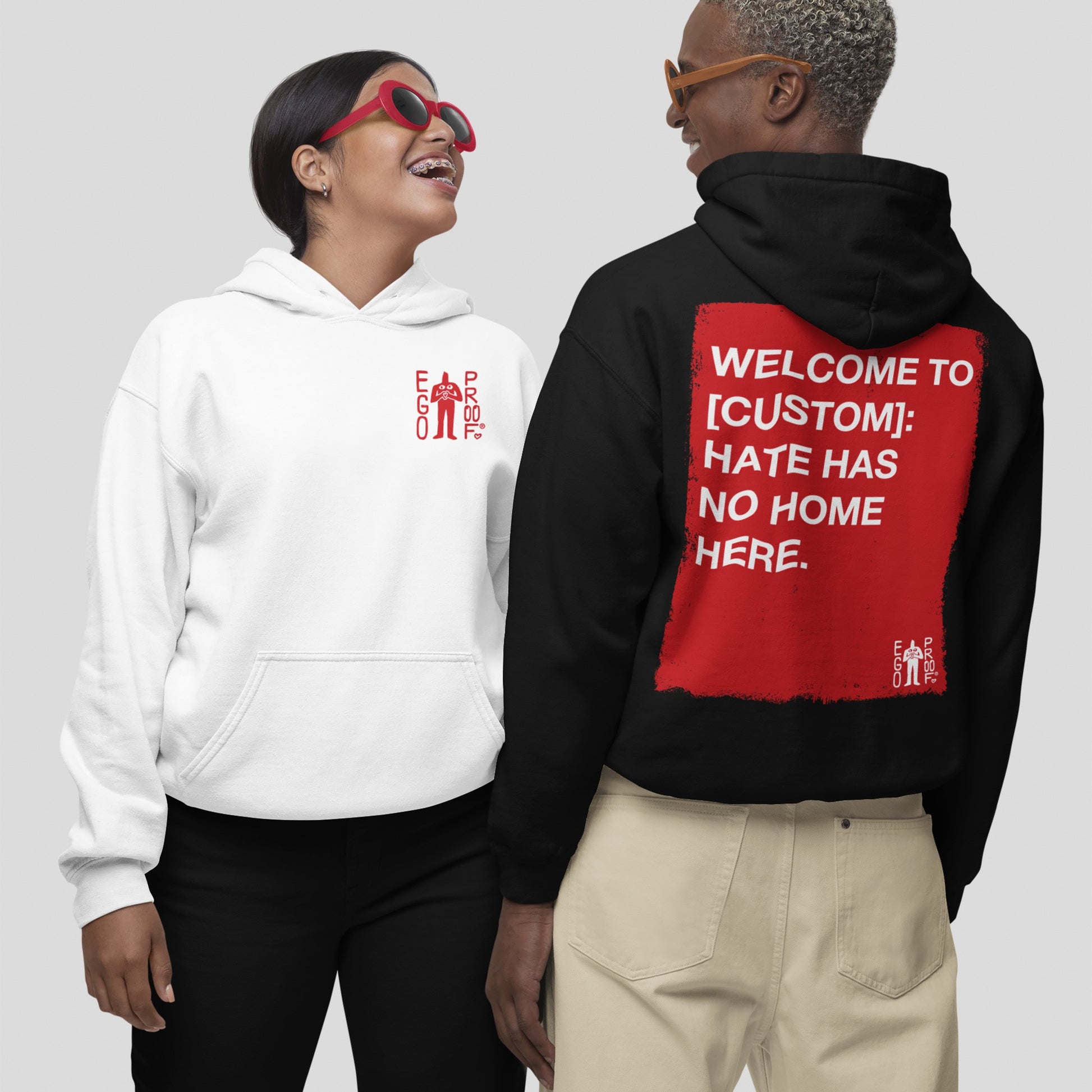 Two models wear the Custom Hate Has No Home Here Hoodie. The left features a red EGO PROD. logo, while the right showcases a bold red square with an inclusive message on the back.