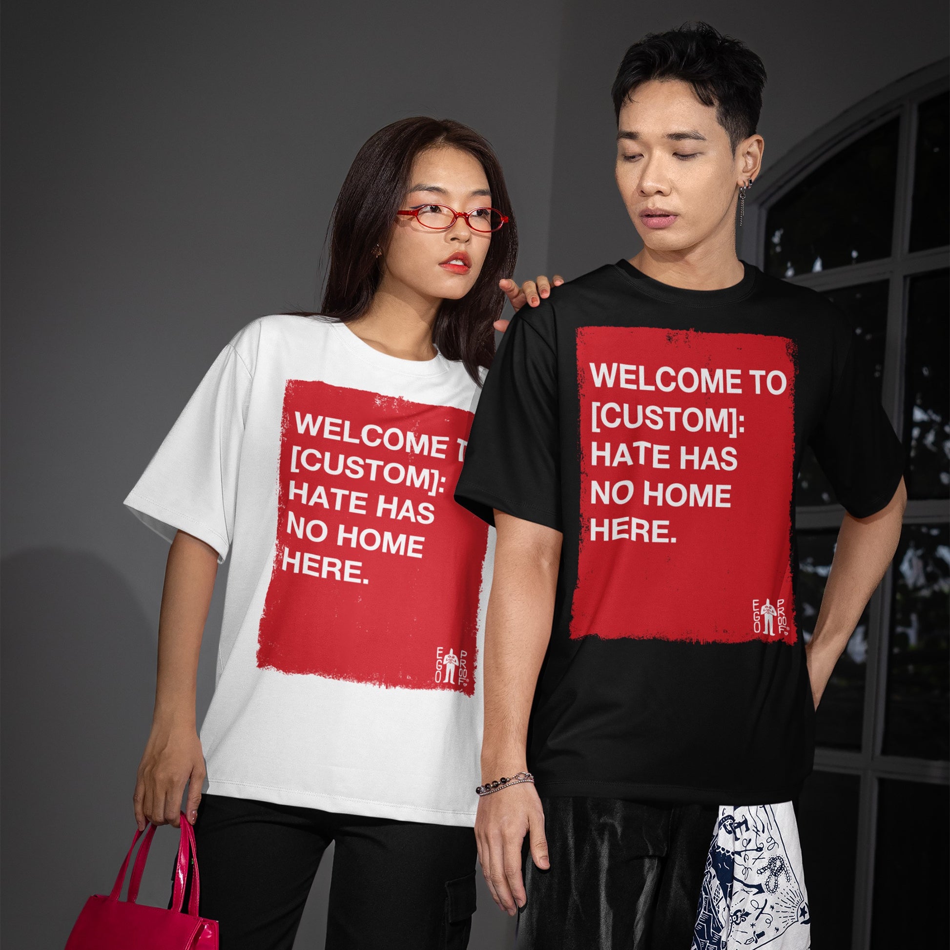 Two young adults wear Custom Hate Has No Home Here T-shirts featuring a bold, red square with a customizable anti-hate message, styled with patterned shorts and a pink tote.