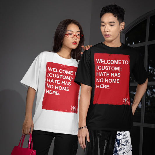 Two young adults wear Custom Hate Has No Home Here T-shirts featuring a bold, red square with a customizable anti-hate message, styled with patterned shorts and a pink tote.