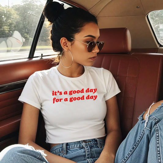 Good Day Women's Baby Tee: A young woman in a car wears this cropped T-shirt, printed with it's a good day for a good day, paired with light-wash jeans, hoop earrings, and sunglasses.