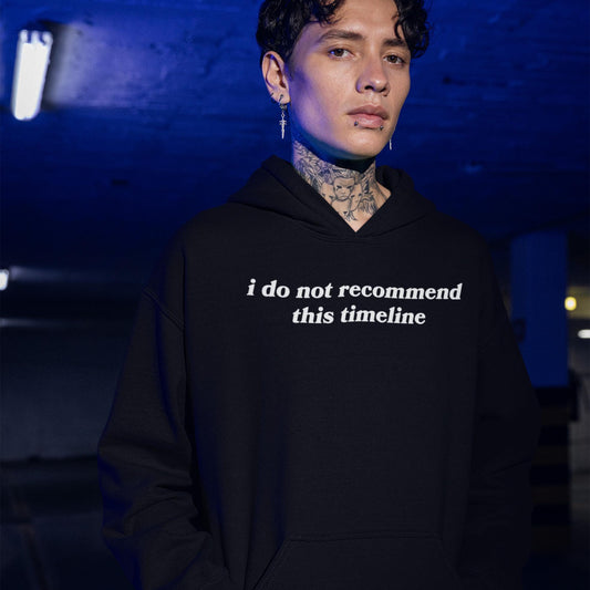 Model wearing black Timeline Hoodie with white text "i do not recommend this timeline" in urban dimly lit setting.