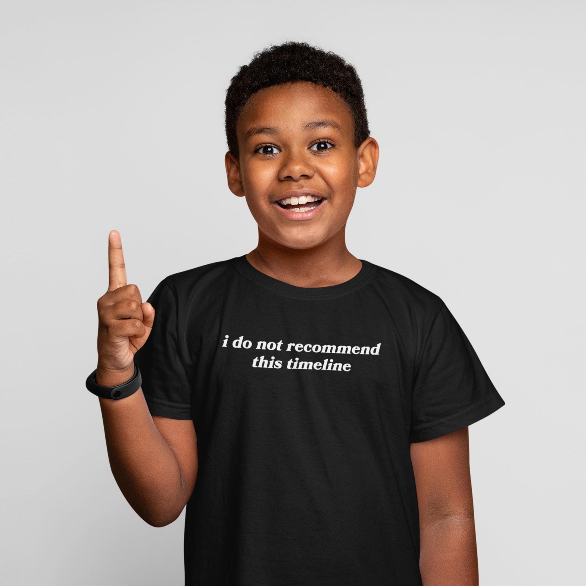 Young boy smiling, wearing the Timeline Kids T-shirt EU featuring the phrase “i do not recommend this timeline” in white text, paired with a black fitness tracker on his left wrist.