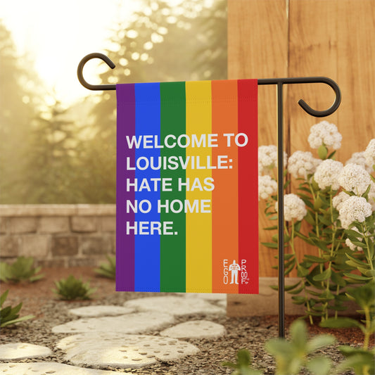 Custom Hate Has No Home Here Pride Garden Flag displayed in a landscaped yard, mounted on a black metal stand, showcasing vibrant rainbow stripes and a welcoming message. Logo reads EGO PROOF.