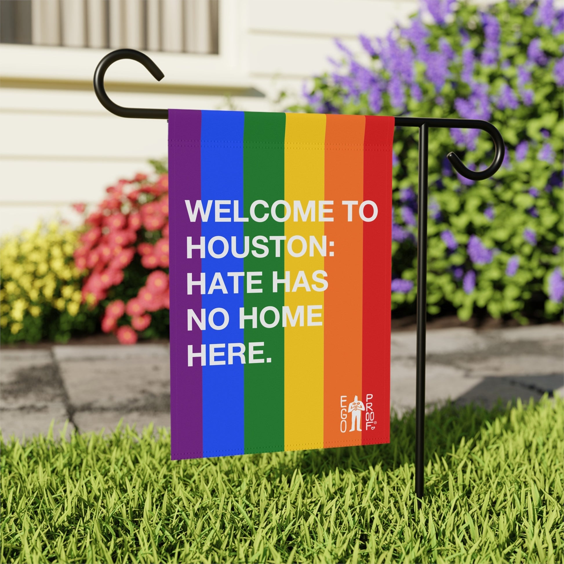 Custom Hate Has No Home Here Pride Garden Flag features vibrant rainbow stripes and bold white text, mounted on a black metal stake in a garden, symbolizing inclusion and love.