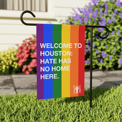 Custom Hate Has No Home Here Pride Garden Flag features vibrant rainbow stripes and bold white text, mounted on a black metal stake in a garden, symbolizing inclusion and love.