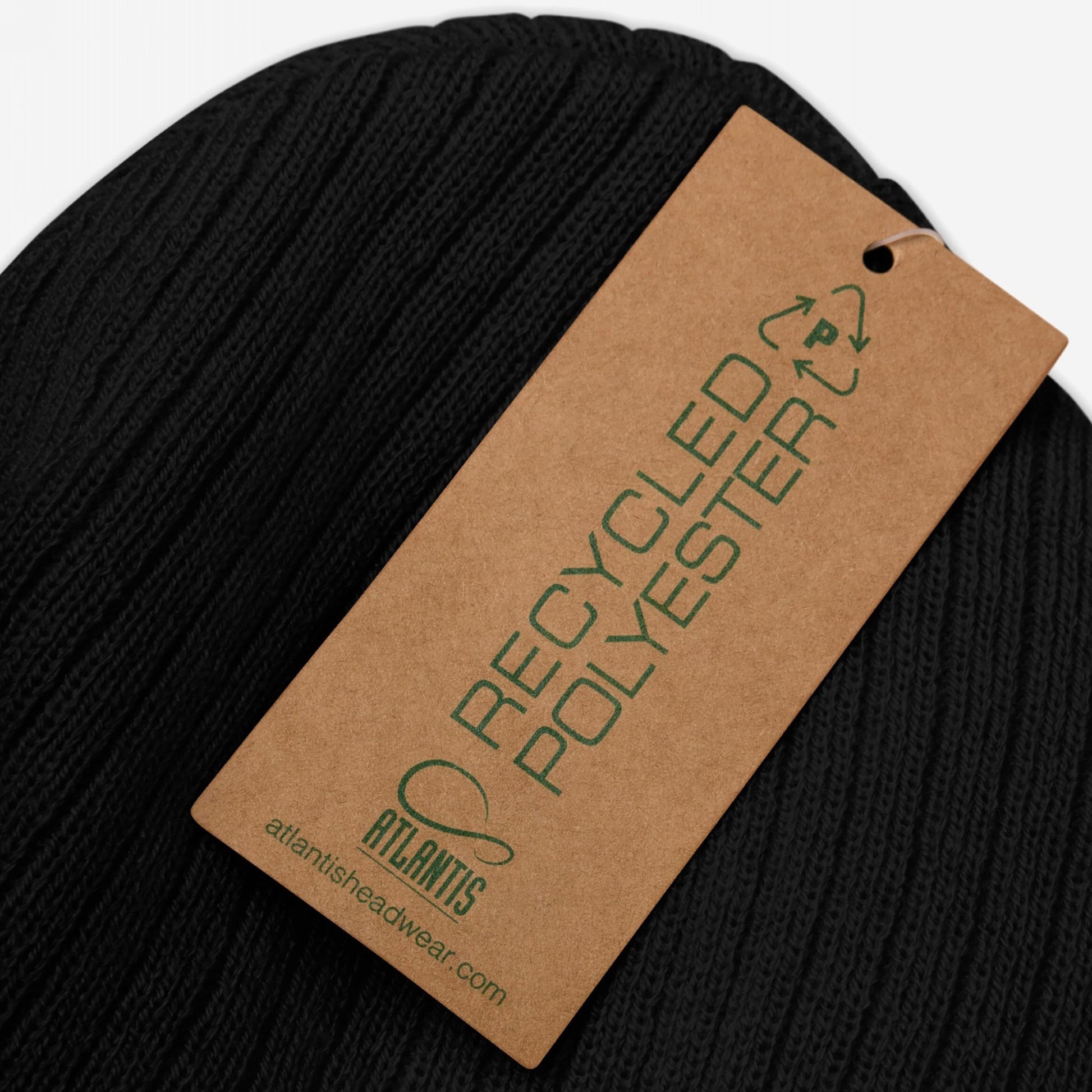 Close-up of a brown hang-tag on black knit fabric, stamped with 'RECYCLED POLYESTER' and 'ATLANTIS.' Represents the 'Make Art Every Day Hat,' designed to inspire creativity with embroidered messaging.