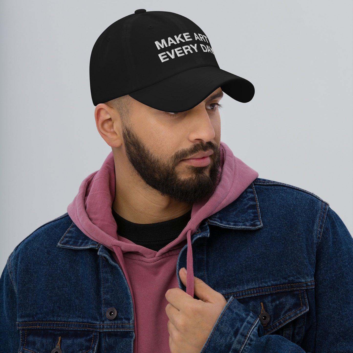 A young man wears the Make Art Every Day Hat, a black snapback with bold embroidery. He's styled in a pink hoodie and denim jacket, embodying Egoproof's creative, resilient spirit.