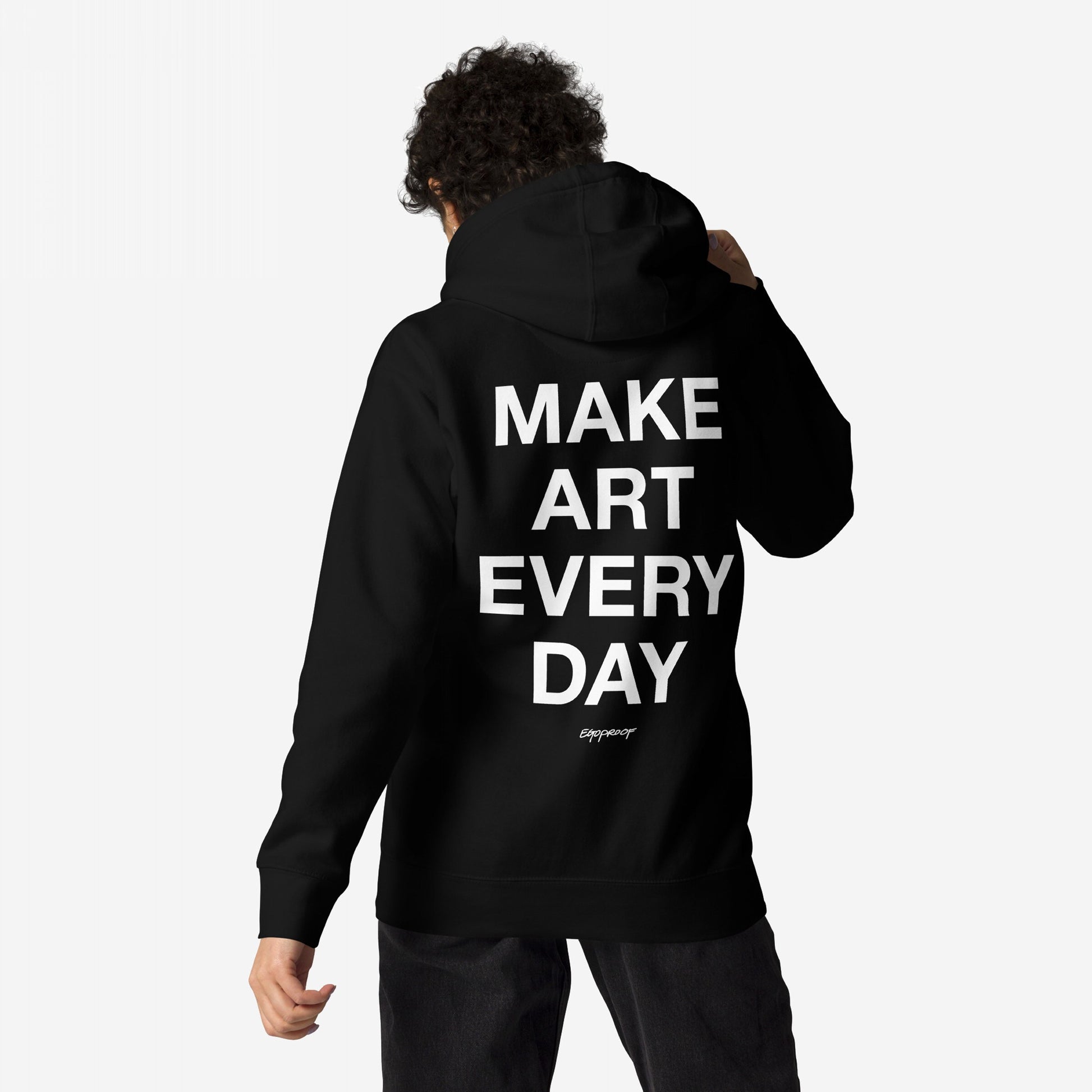 Person wearing 'Make Art Every Day Hoodie EU' with bold white lettering on the back. Features dark curly hair, set against a light background, embodying creativity and daily artistic inspiration.