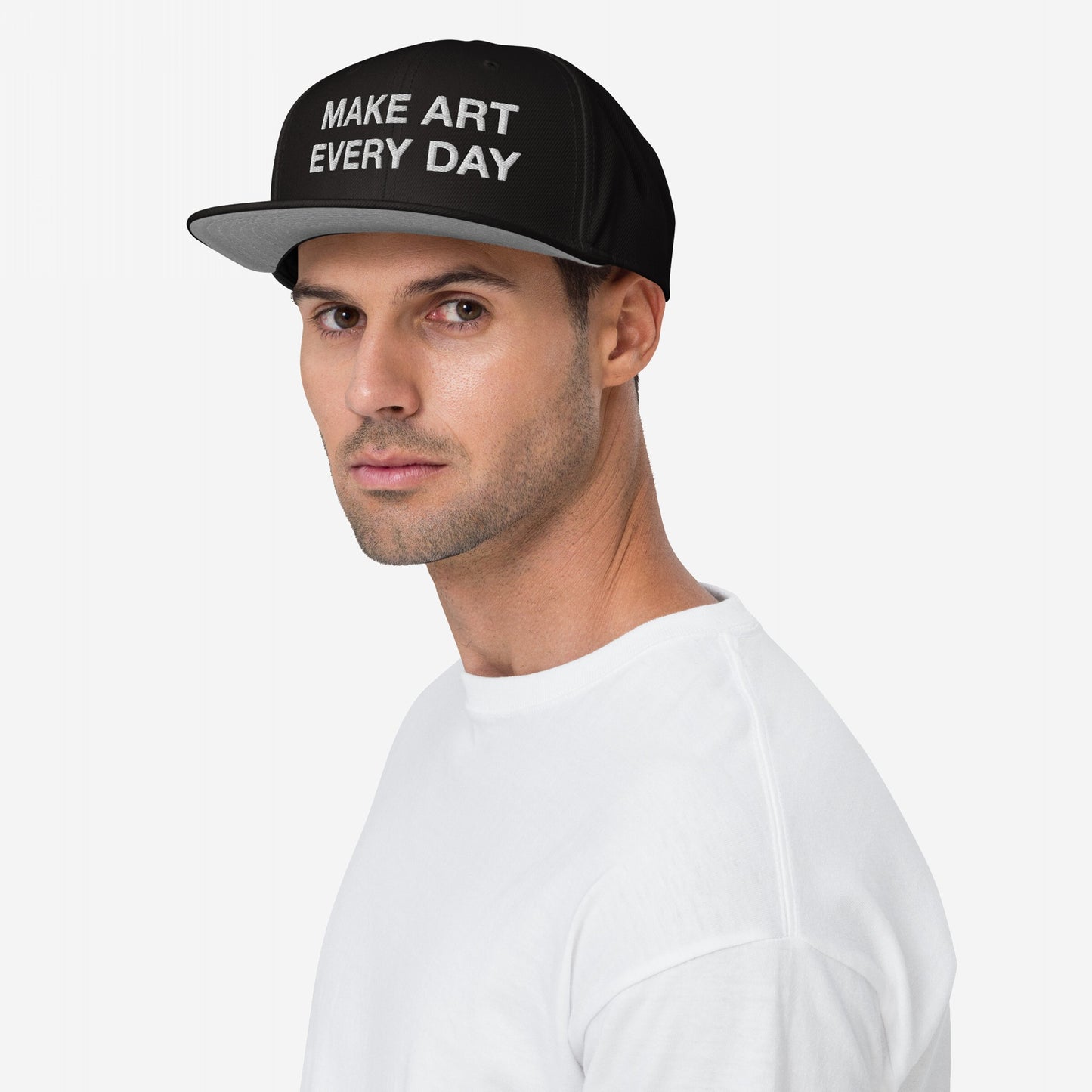 Young man wearing the Make Art Every Day Hat, a black flat-brimmed cap with white embroidered slogan, exemplifying creativity. Designed for adjustable fit, ideal for artistic inspiration and everyday wear.
