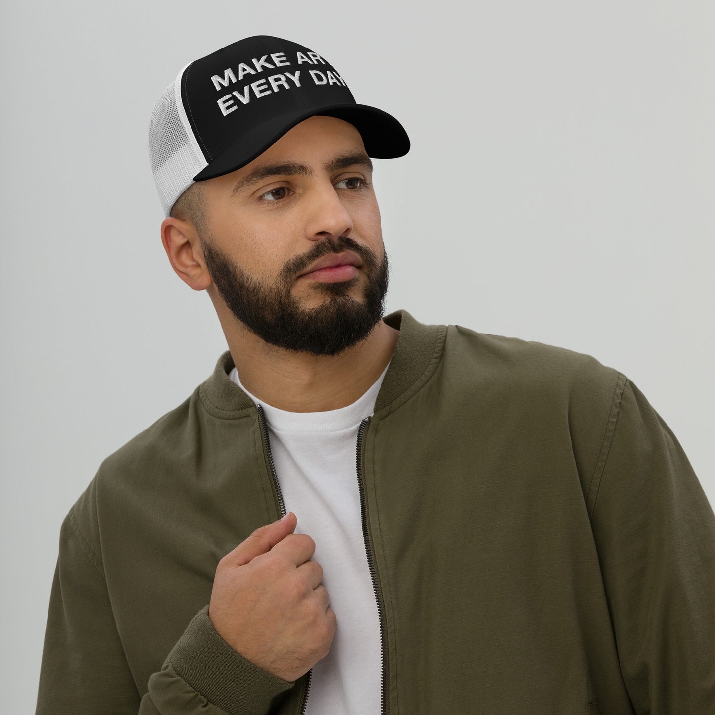 A bearded man wearing the Make Art Every Day Hat, a black-and-white trucker cap with impactful embroidery, styled with an olive jacket over a white T-shirt, embodying creativity and perseverance.