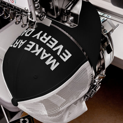 Make Art Every Day Hat being embroidered on an industrial machine, showcasing bold white lettering on the front panel of a black-and-white mesh trucker cap.