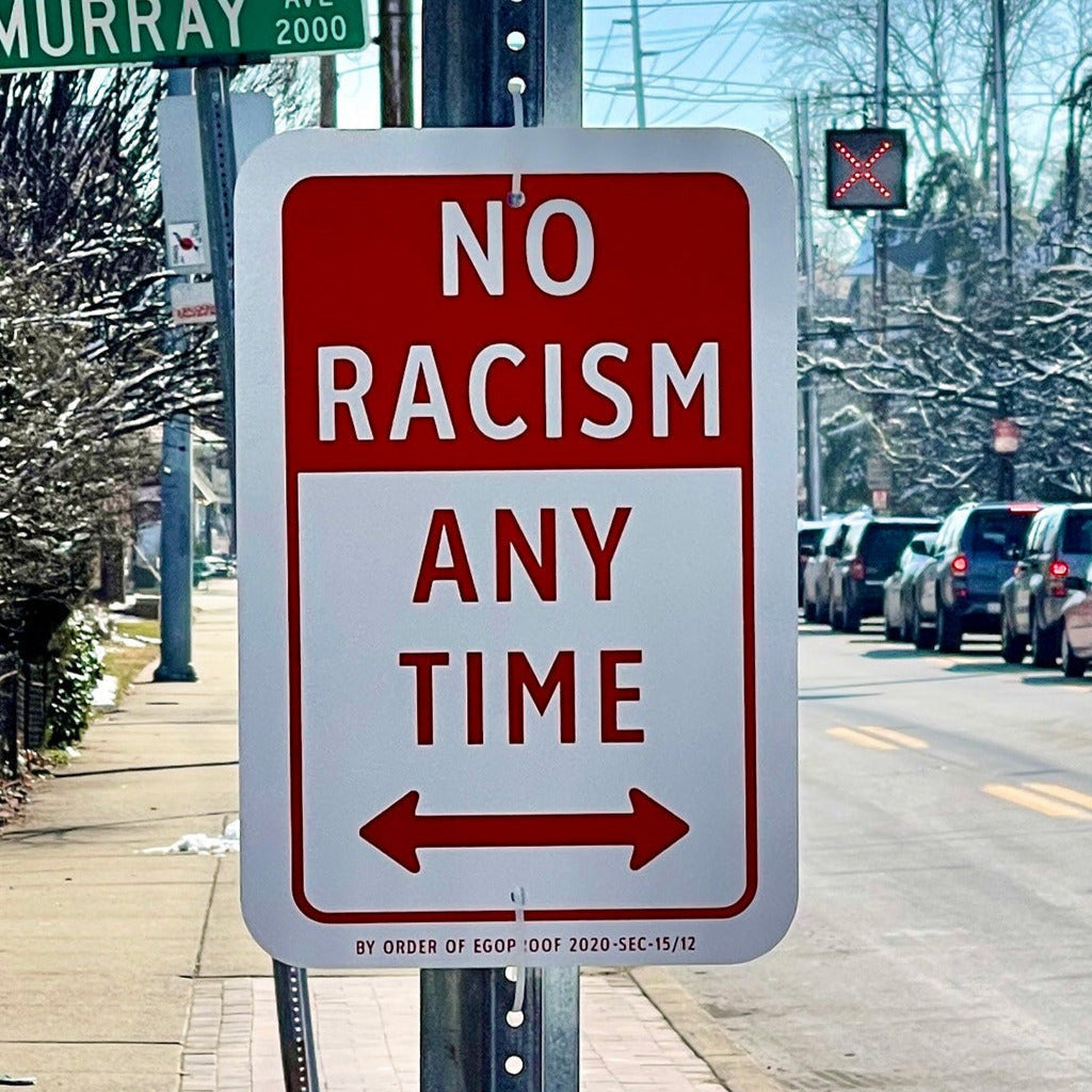 Street sign with 'No Racism Any Time' on a pole, with cars and trees in the background.