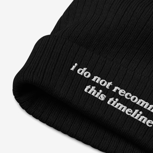 Close-up of the Timeline Hat, a black ribbed knit beanie with a folded cuff, featuring partial white embroidery: “i do not recommend this timeline.” Ideal for sarcastic flair amidst modern chaos.